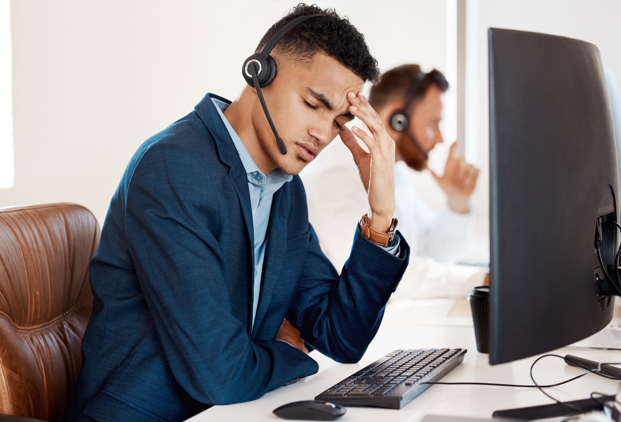 A support agent at his workstation, headset on, head in his hands, mid-shift burnout.